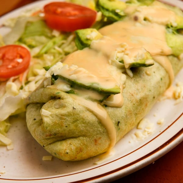 A large burrito covered in cheese sauce and avocado slices, served with a side salad.