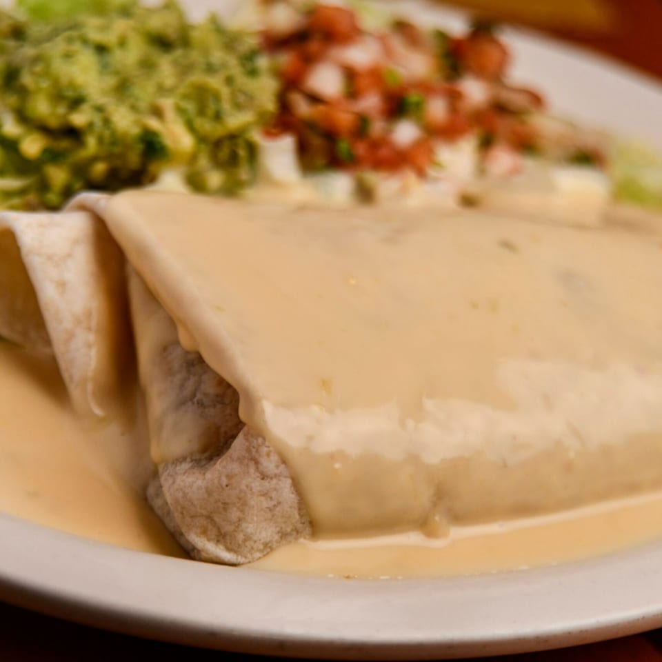 A cheese-covered burrito with guacamole and pico de gallo on a white plate at Blue Agave.